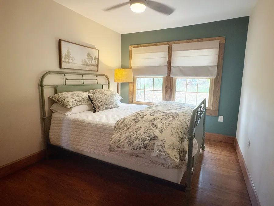 A bedroom with a pale iron bed against a deep green wall, twin windows alongside