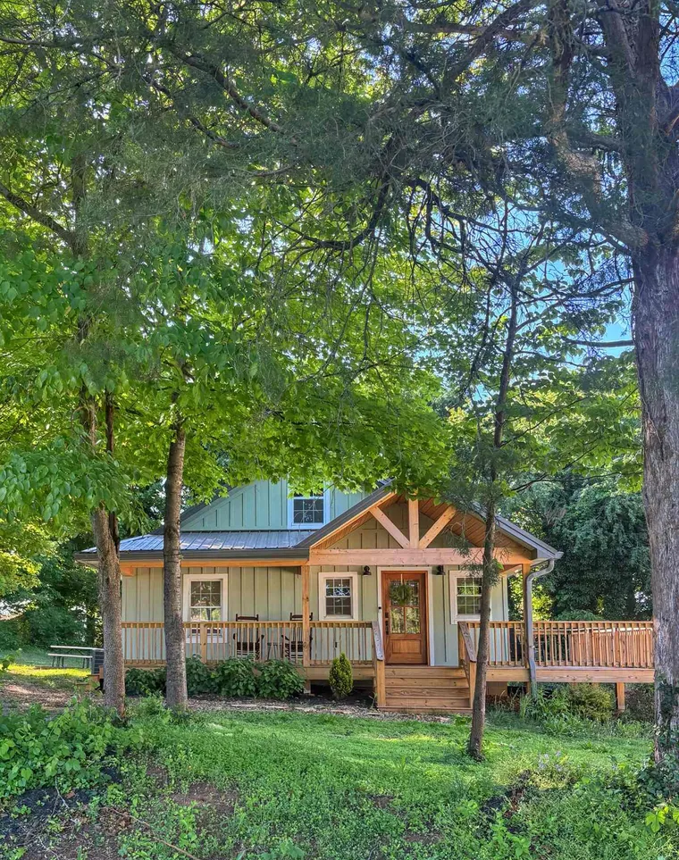 The pale green cottage with its timber-framed porch, framed by tall leafy trees in summer
